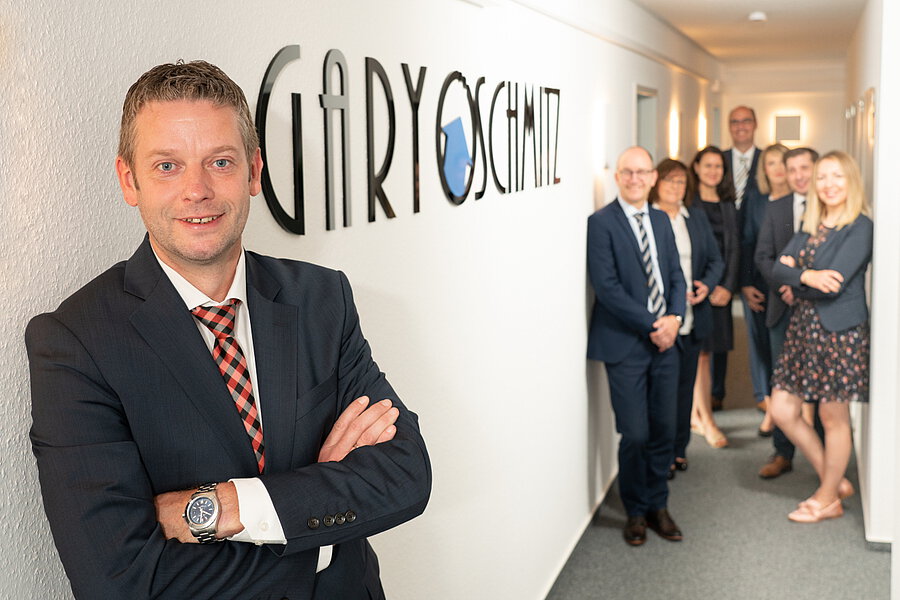 Gary Schmitz vor dem Firmenschriftzug im Büroflur, dahinter sein Team